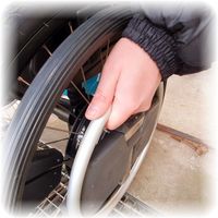 Detailaufnahme eines Rollstuhlhinterrades mit Wheeldrive Antrieb. Die gezeigte Hand am Greifreifen zeigt wie es aussieht, wenn man den Rollstuhl nach vorne fahren möchte.